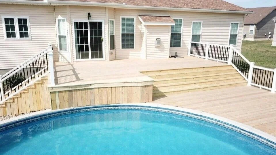 Backyard with a wooden deck, stairs leading down to a swimming pool, and a house with light-colored siding, multiple windows, and a sliding glass door.