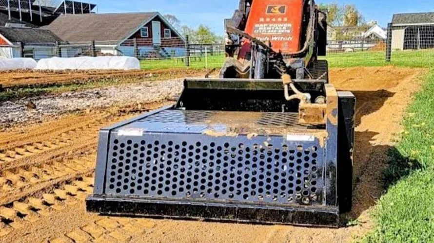 A compact excavator with a grading attachment working on a dirt path in a rural yard with house and fencing in the background.