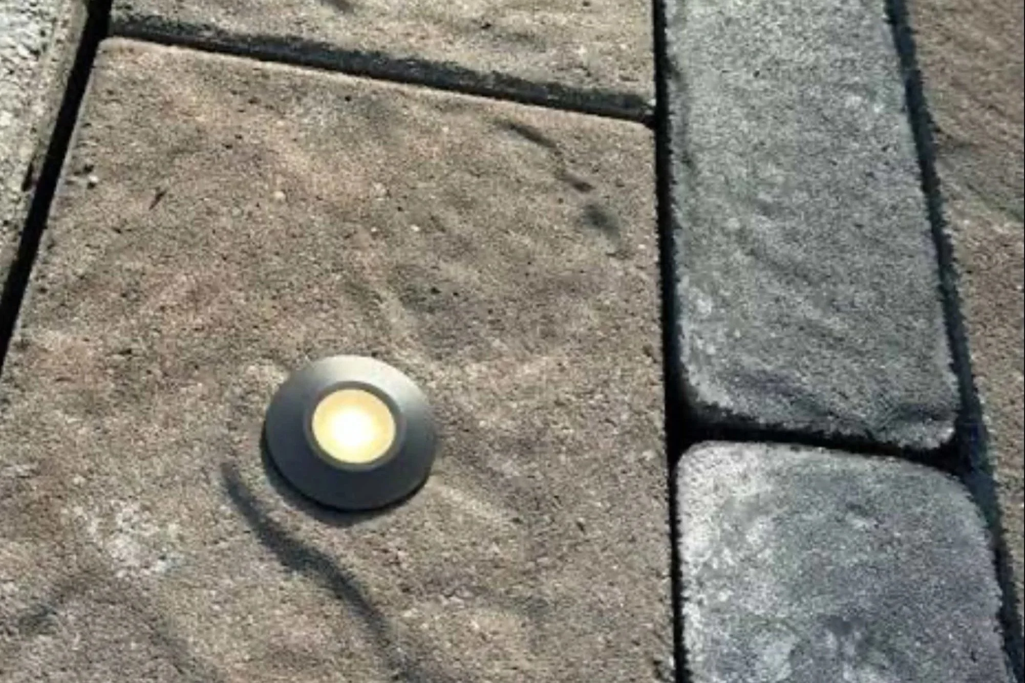 Close-up of a landscaped area with a circular ground light, sand, and stone borders.