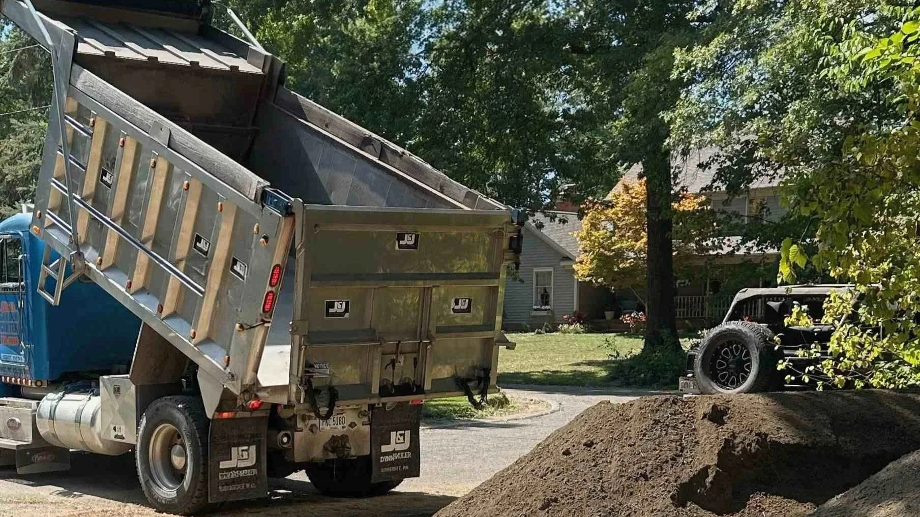 A truck with an open dump bed loaded with dirt parked in a residential neighborhood. A pile of dirt is in the foreground, and a black off-road vehicle is partially visible behind trees on the right.