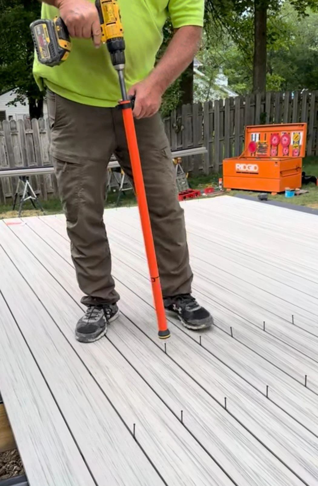 A person is using a hammer drill to secure deck screws into a new outdoor patio deck in a backyard with a wooden fence and trees. A toolbox with tools and supplies is visible in the background.