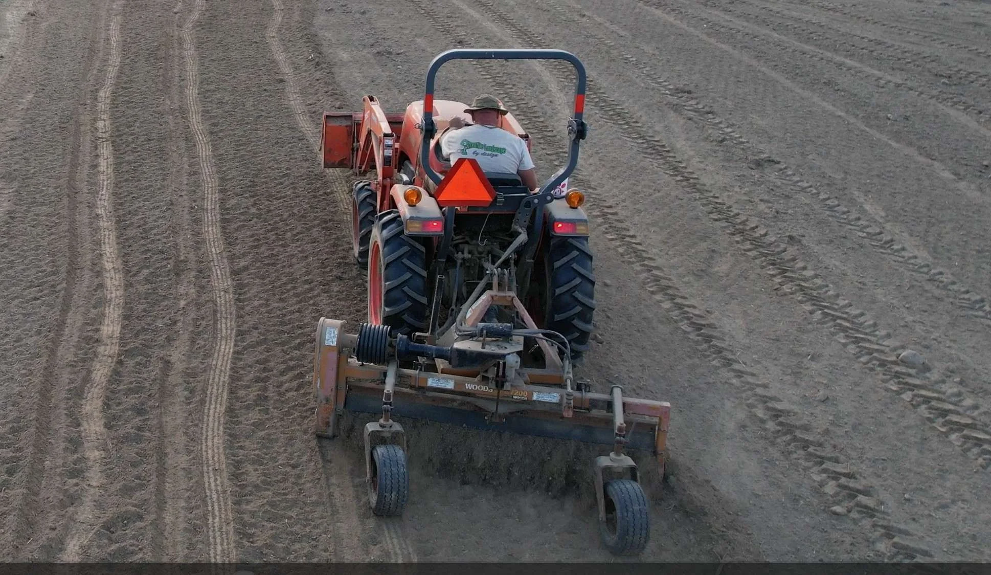A person operating a tractor on a large field, preparing the soil with a tiller attached to the back.