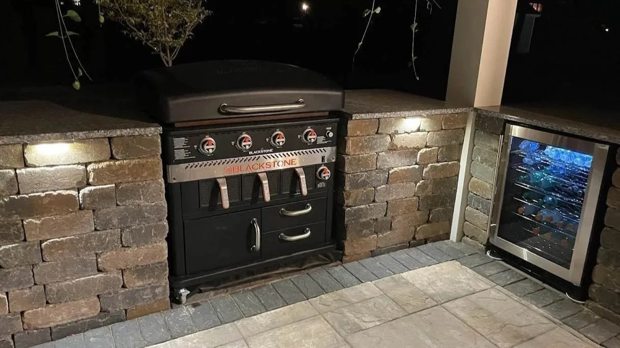 Outdoor area with a black barbecue grill, built into a brick counter, next to a small wine cooler filled with bottles.