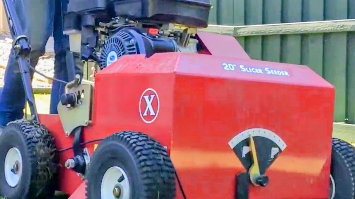 Red rectangular machine with a black motor on top, four large tires, and a gauge on the front, located outdoors next to a green wooden fence.