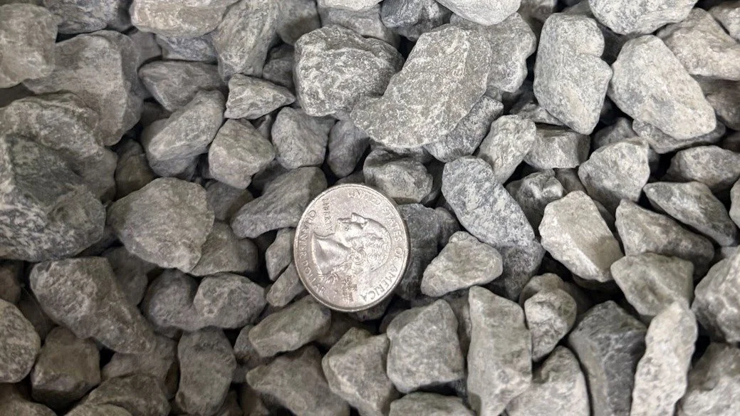 A close-up of gravel stones with a quarter coin for size comparison, showing various gray rocks.