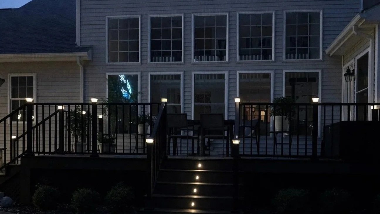 A backyard deck attached to a house at night, with small lights on the railing, chairs, and potted plants