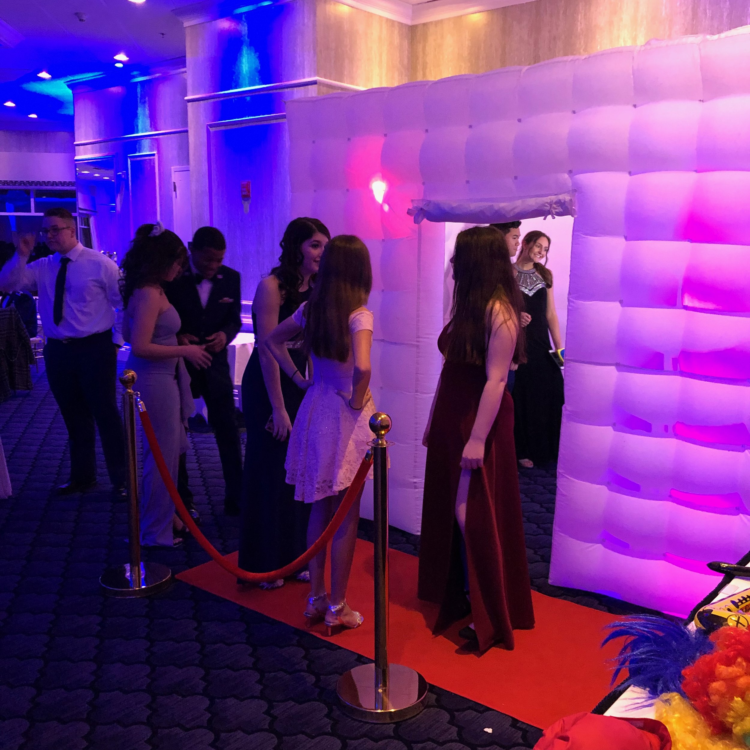 Party guests waiting beside a white inflatable photo booth enclosure lit in purple and pink.
