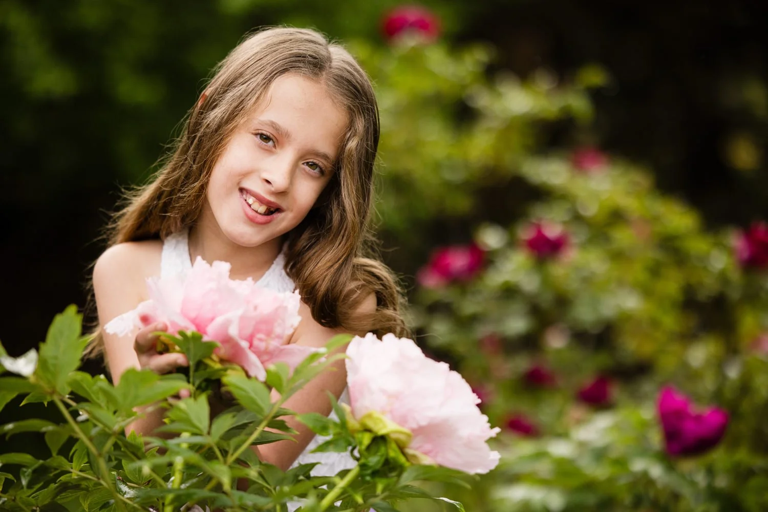 A young girl with long brown hair and a girl smile holding pink flowers in a garden with green foliage and pink flowers in the background at Webster Arboretum