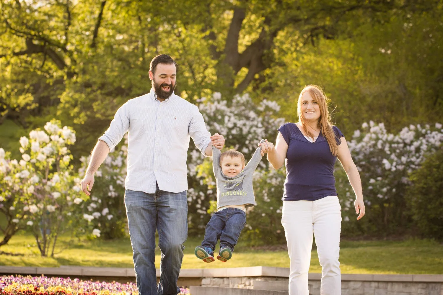 A family of three, a man, woman, and young boy, enjoying a walk in Highland Park during spring. The boy is being swung by his parents and looks happy in this family portrait.