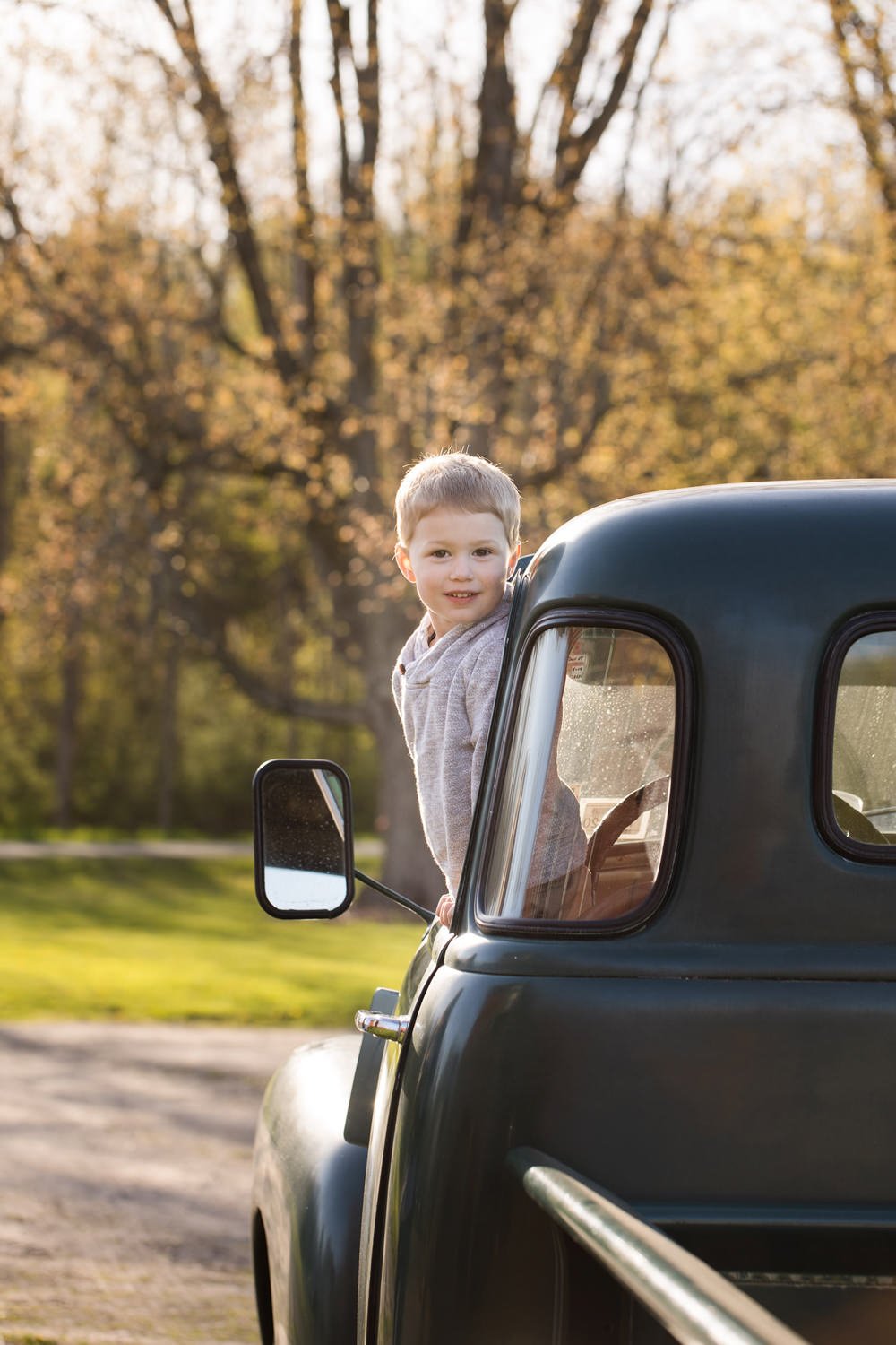 A young boy leaning out of the passenger window of a vintage black truck, outdoors in a park with trees and fall foliage in the background.
