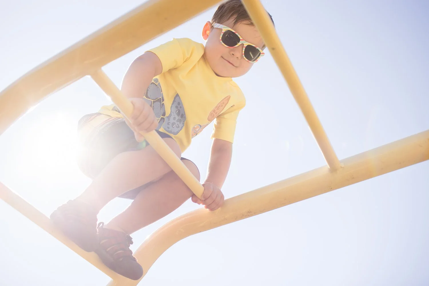 Child wearing sunglasses and a yellow shirt, climbing a yellow playground structure against a bright sky.