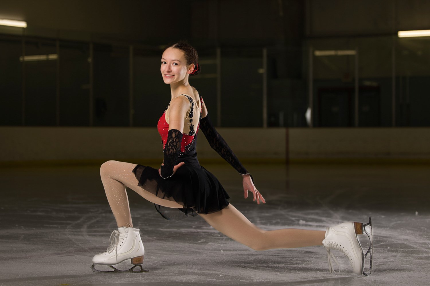 A young woman in figure skates and a black and red skating costume kneeling on an ice rink, smiling at the camera for senior pictures