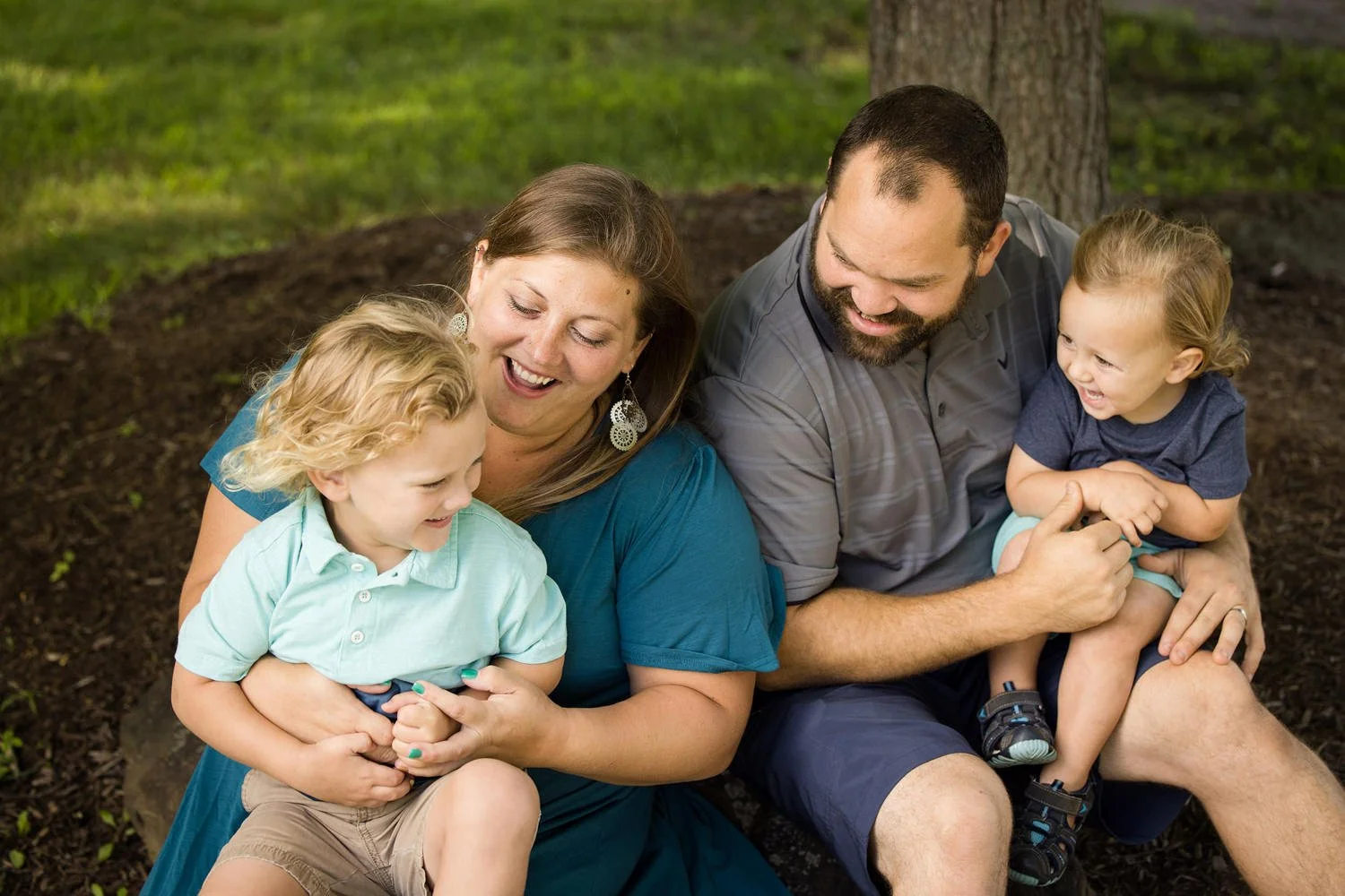 A family of four, including a woman, a man, and two young children, sitting outdoors near a tree, smiling and playing together.