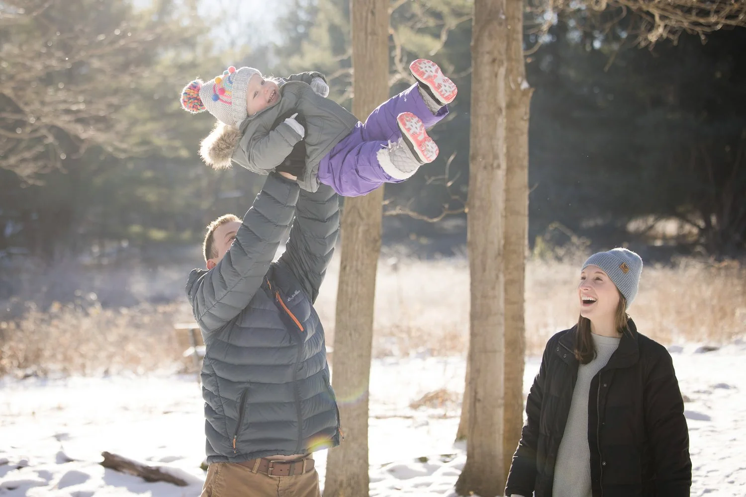 A man and woman playing with a young girl in a snowy Mendon Ponds park during winter. The man lifts the girl in the air while the woman watches and smiles.