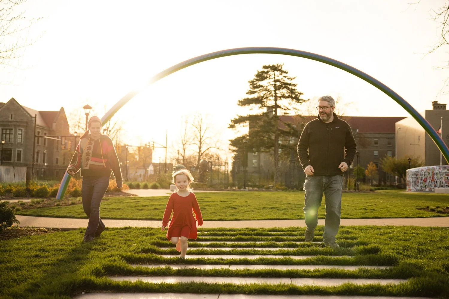 A family of three—an adult man, a woman, and a young girl walking on grass in a park at sunset under a rainbow arch at Memorial Art Gallery in Rochester NY