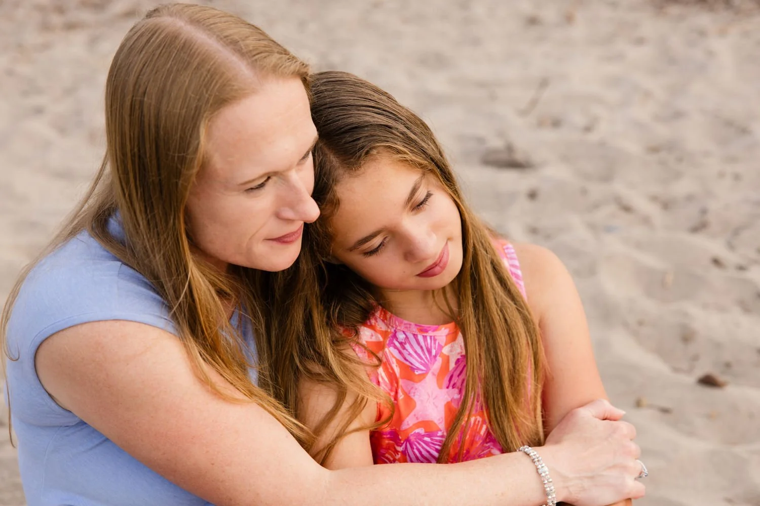 A woman and a girl sitting closely on a sandy beach in Rochester NY, leaning their heads together with eyes closed, sharing a tender moment.