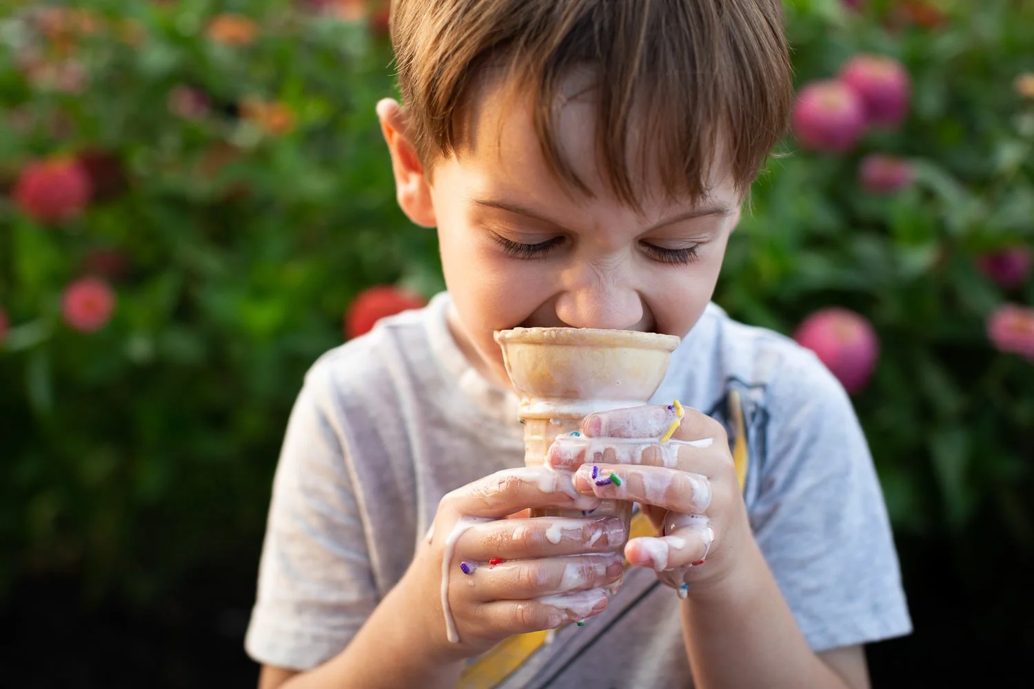 Young boy with messy hands licking an ice cream cone outdoors with pink flowers in the background at a candid family portrait session in Rochester NY