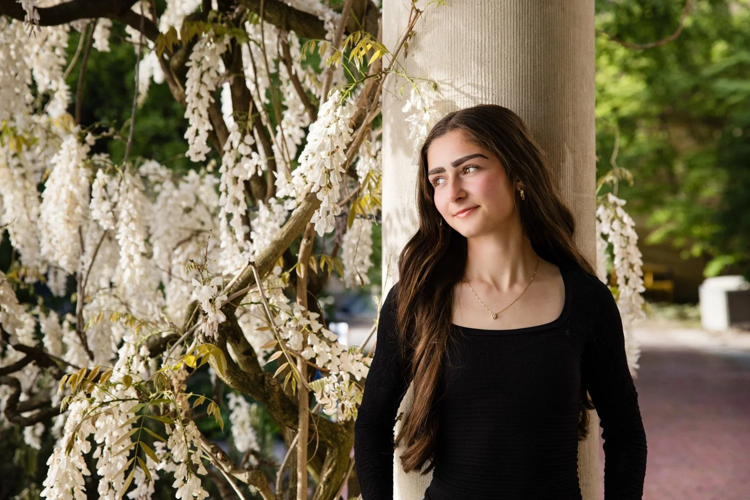 A senior high school girl with long dark hair, wearing a black long-sleeve top and gold jewelry, leans against a beige column at Eastman Museum outdoors among white flowering trees.