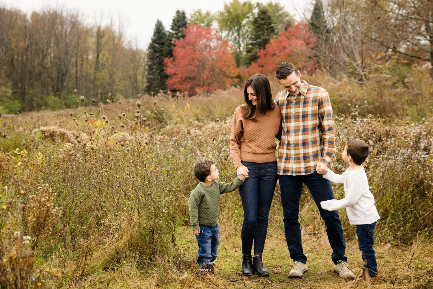 A family of four walking through a fall landscape with trees showing autumn colors, holding hands and smiling at Tinker Park.