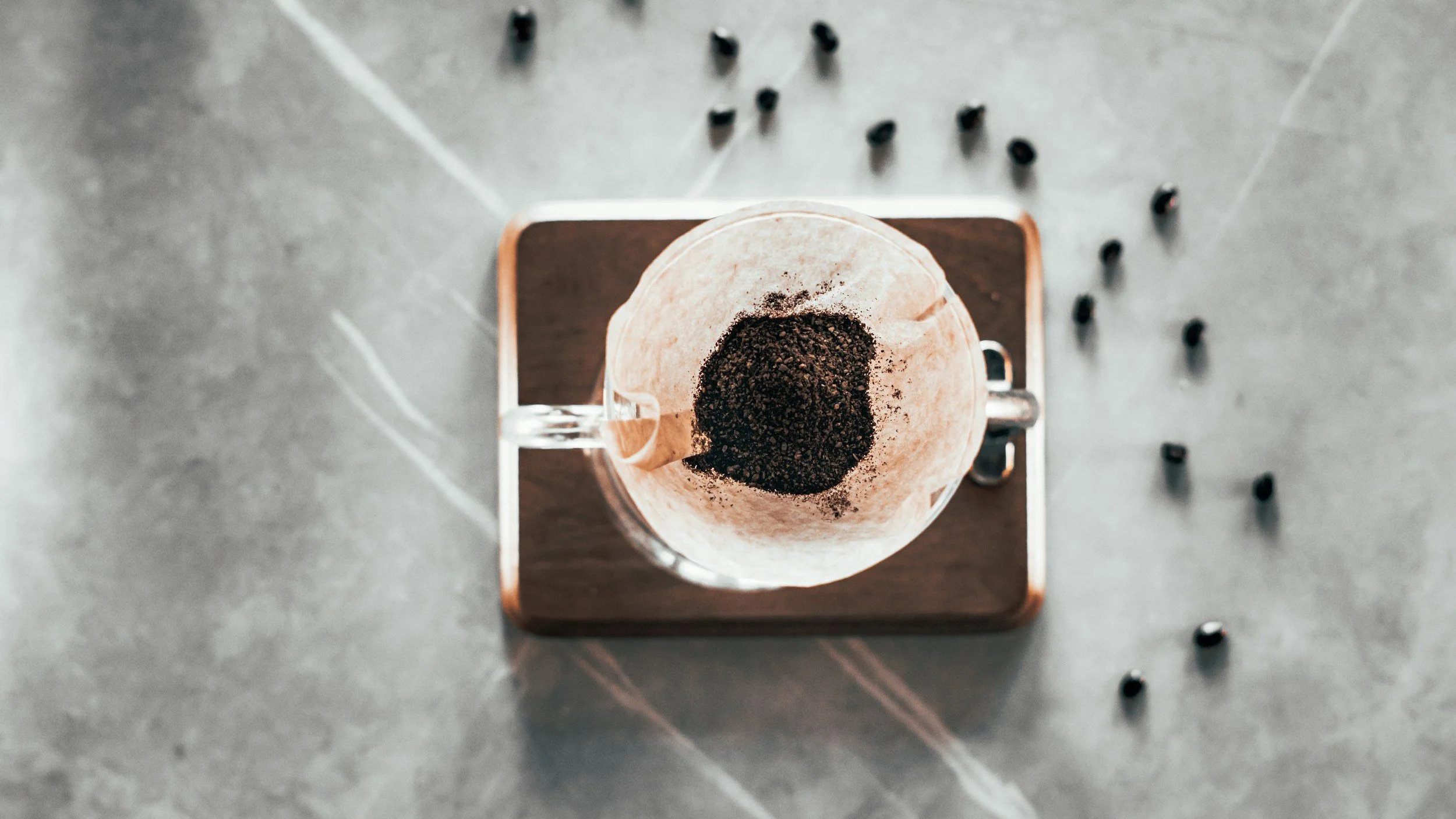 A pour-over coffee setup with a glass mug containing a paper filter filled with coffee grounds, placed on a wooden tray. Scattered around are small black coffee beans on a gray marble surface.
