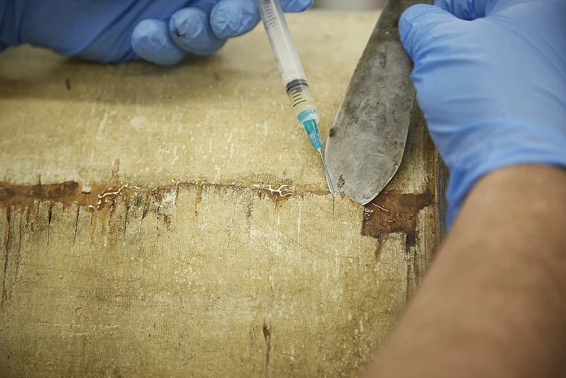 Reversible conservation adhesives are inserted underneath areas of lifting veneer.