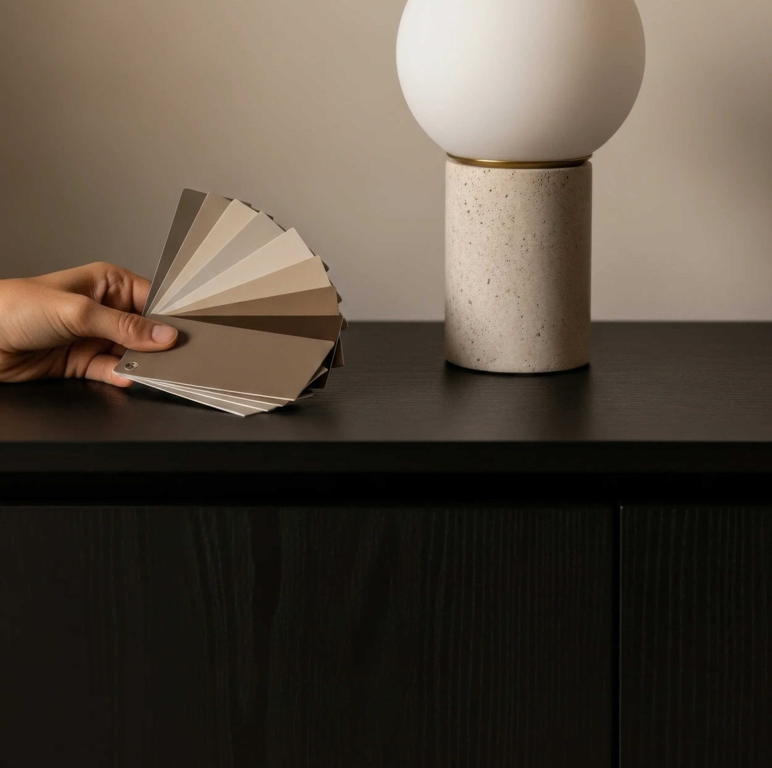 A hand holding a fan of color swatches in shades of beige, cream, and taupe in front of a minimalist lamp with a spherical white shade and a cylindrical textured base on a dark surface.