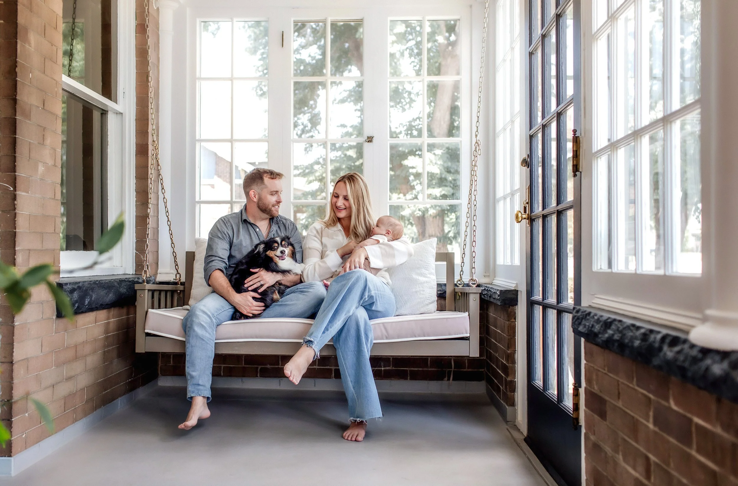 Denver Lifestyle Newborn Session at home with infant and dog on a porch swing with new parents - mom holding baby and dad holding the dog looking at each other and smiling