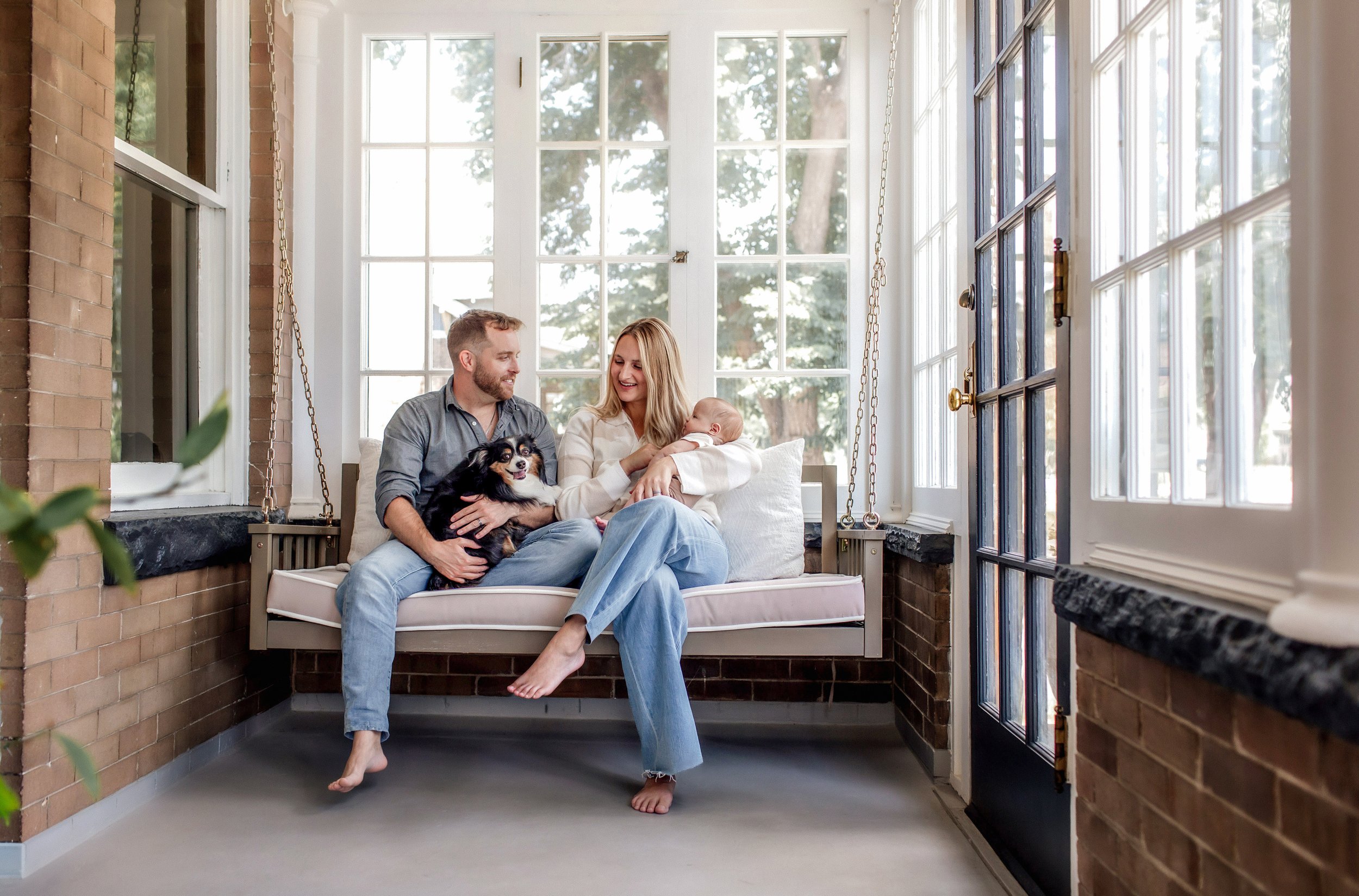 Denver Lifestyle Newborn Session at home with infant and dog on a porch swing with new parents - mom holding baby and dad holding the dog looking at each other and smiling