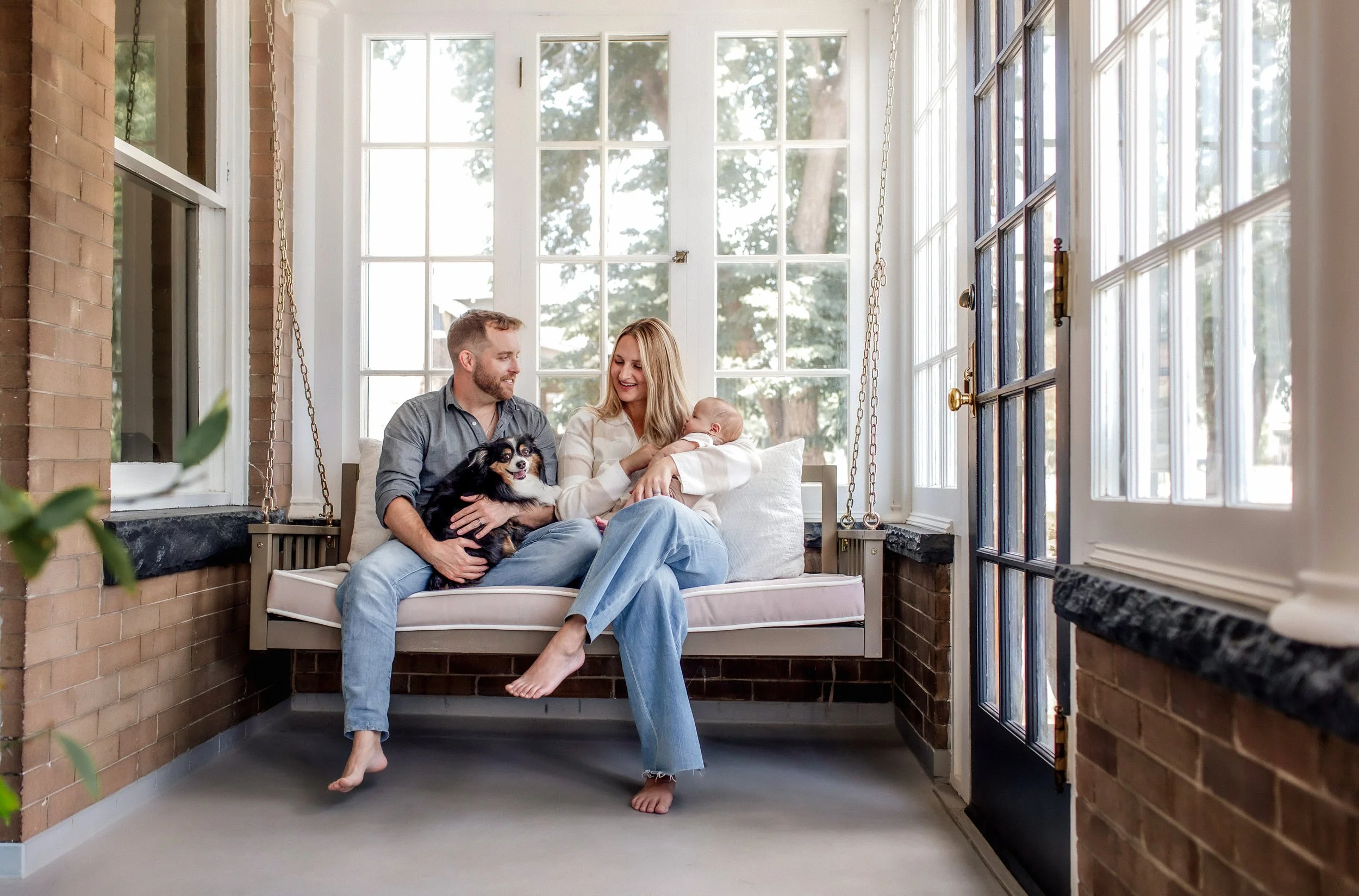 Denver Lifestyle Newborn Session at home with infant and dog on a porch swing with new parents - mom holding baby and dad holding the dog looking at each other and smiling
