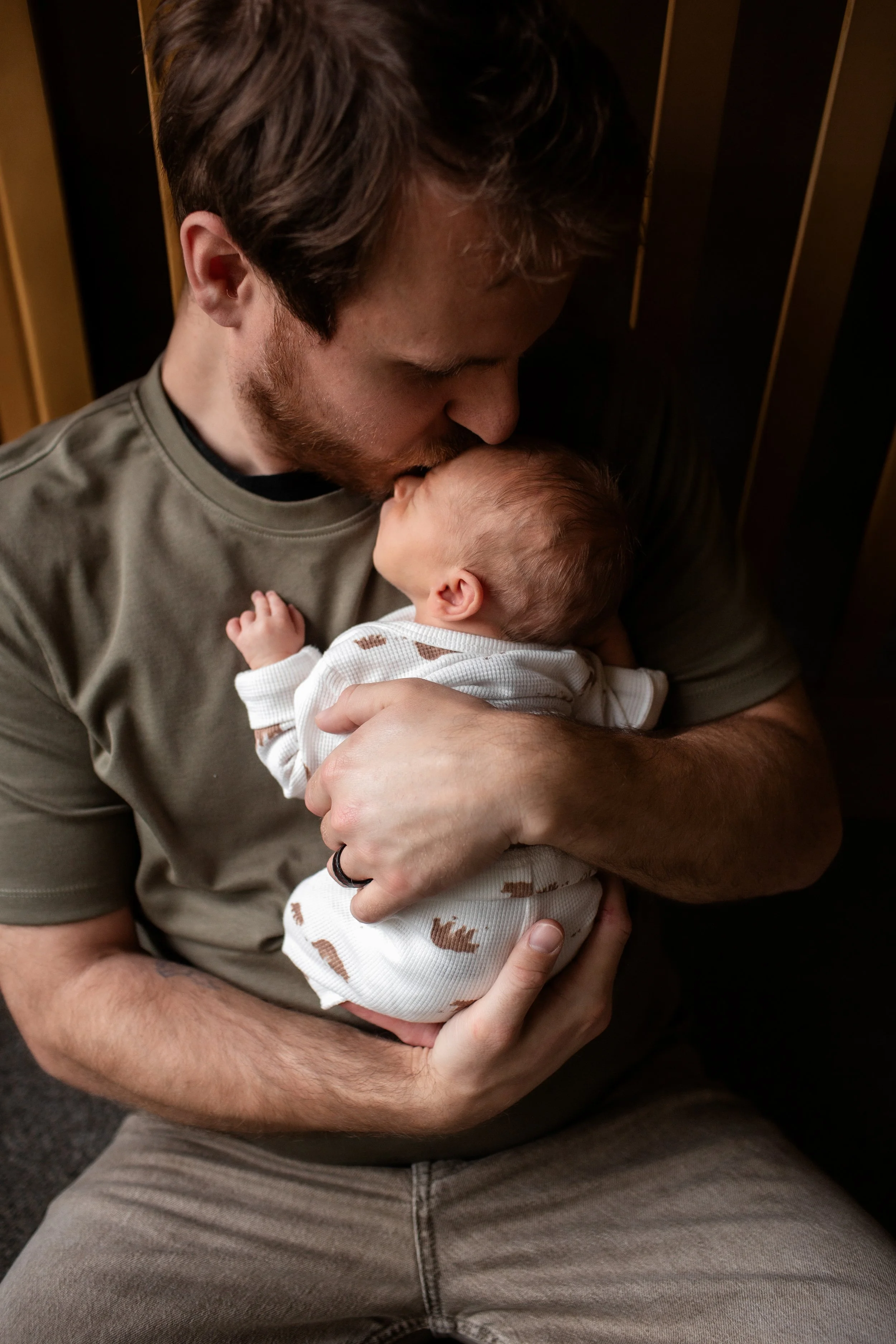 Denver Newborn Lifestyle photos in home with infant and parents Dad sitting in front of crib on floor kissing baby on the forehead