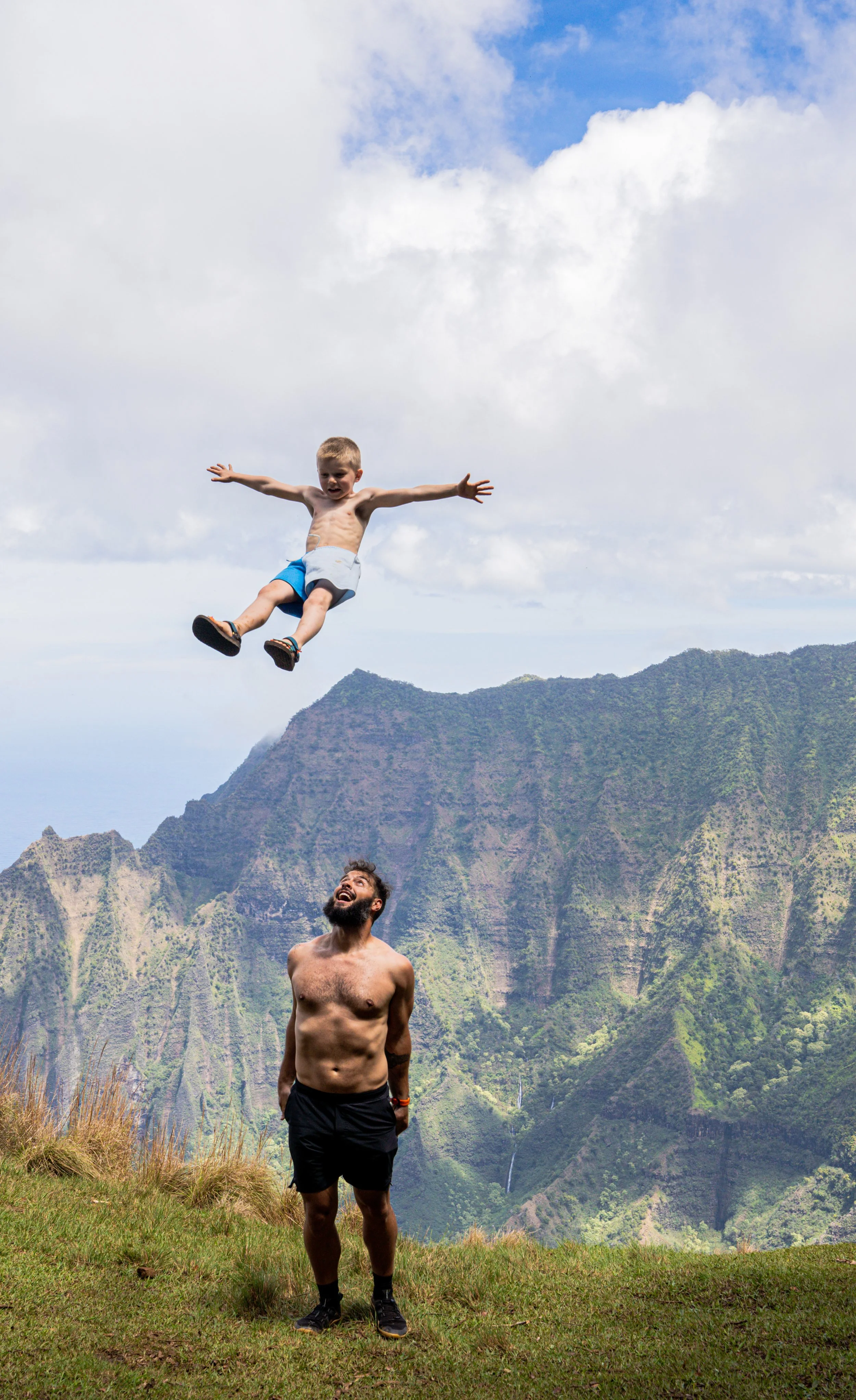 Mike Aidala tossing a young boy into the air on a cliff in Kauai Hawaii with the Napali Coast in the background
