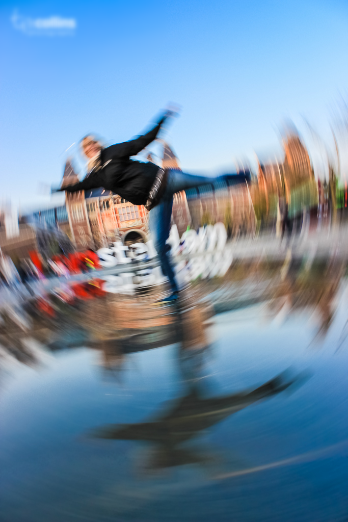 Blurred motion photo of a person skating near a canal with historic buildings in the background, under a clear blue sky.