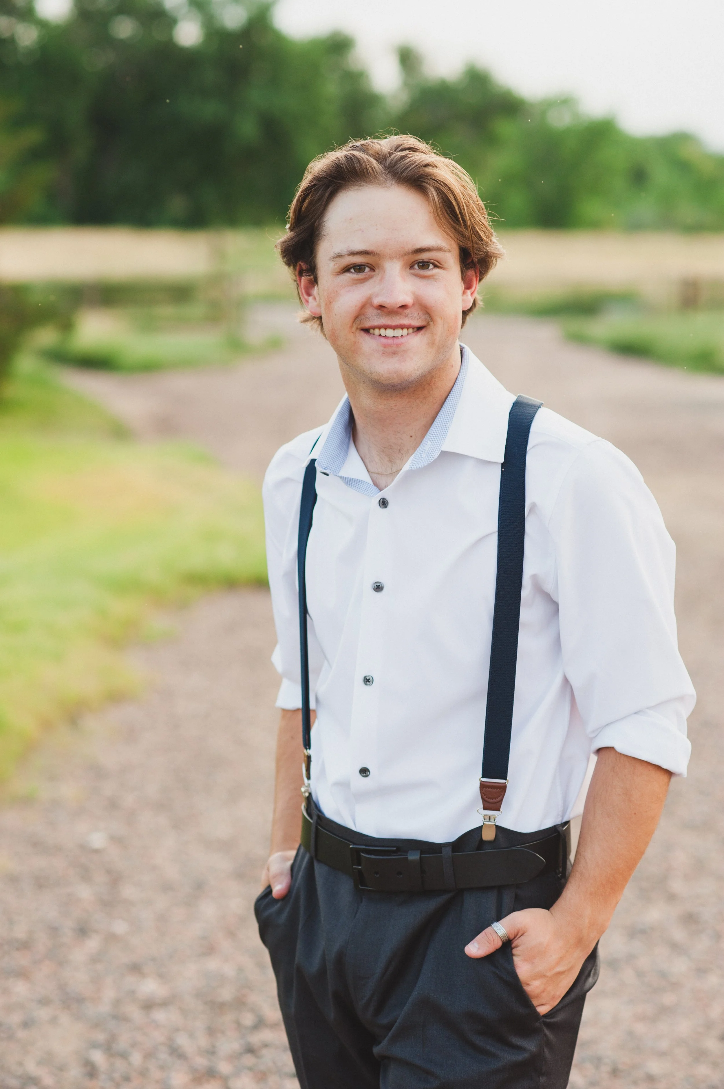 A young man high school senior with brown hair wearing a white button-up shirt, black suspenders, and dark pants, standing on a gravel path outdoors with green trees and fields in the background, at 17 mile house park in Parker CO