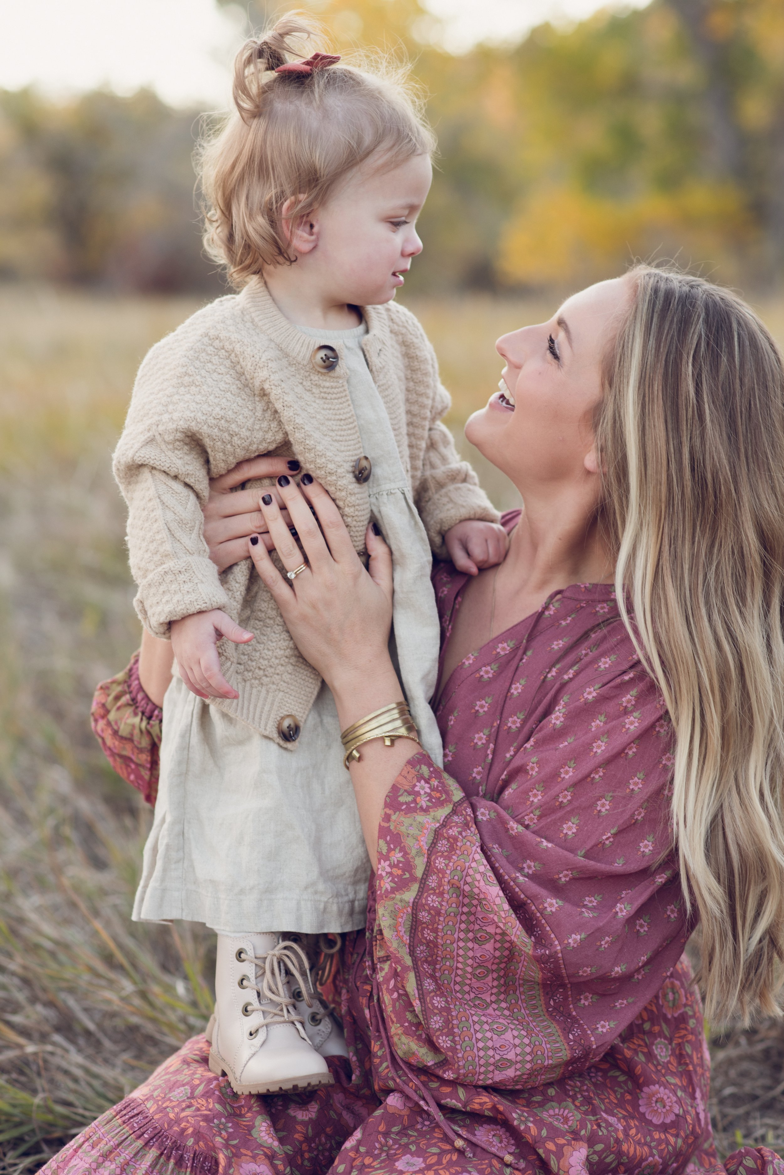 A woman holding and smiling at a young girl in an outdoor setting with fall foliage in the Denver Metro Area near the foothills for family photos.