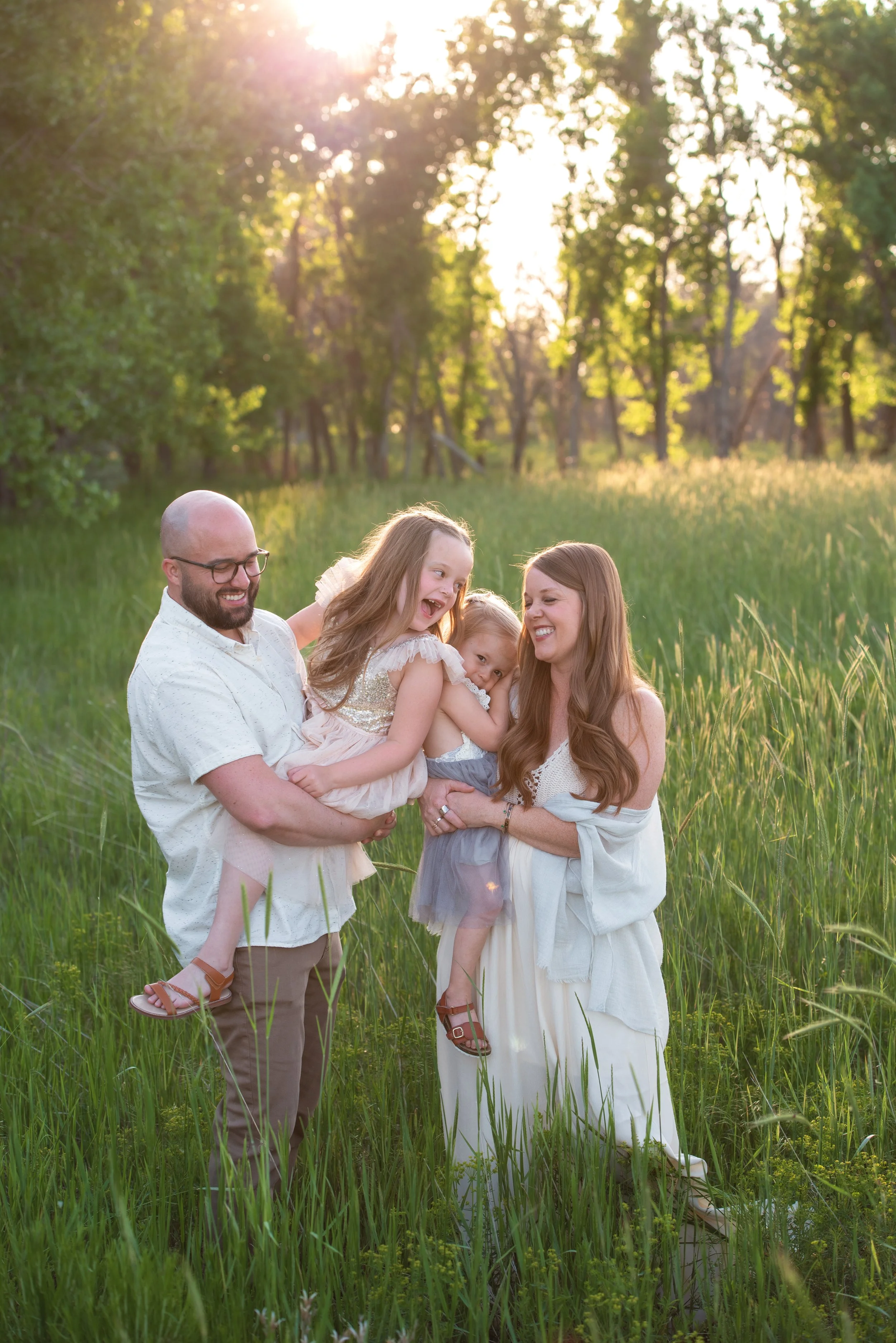 A family of five, including two young girls, a woman, and a man holding an infant, playing and holding hands in a grassy field with mountains in the background.