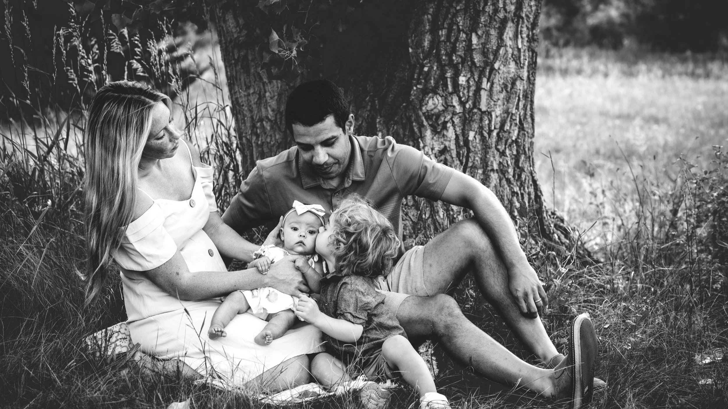 Denver Top Photographer captures A family of four sitting on the grass near a tree in CHerry Hills Village, with mother, father, and two young children, one girl and one baby, outdoors for family photos