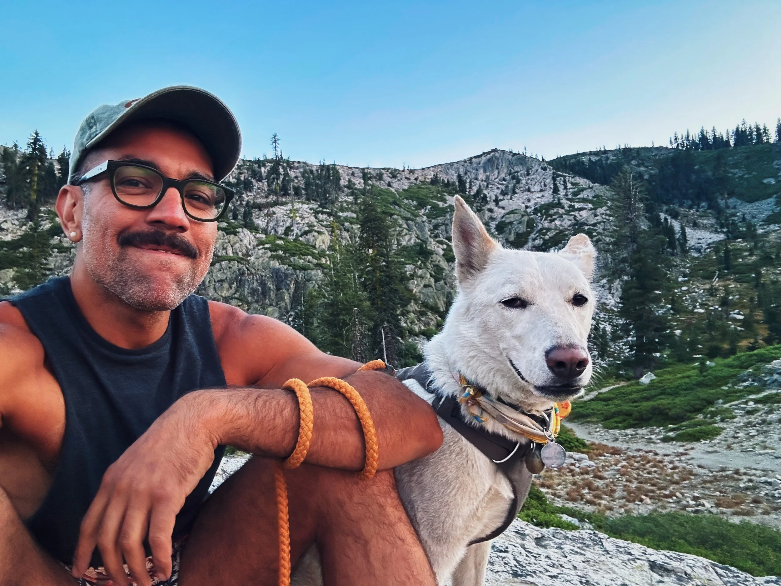 A man with glasses and a cap taking a selfie with a white dog in a mountainous outdoor setting during daytime.