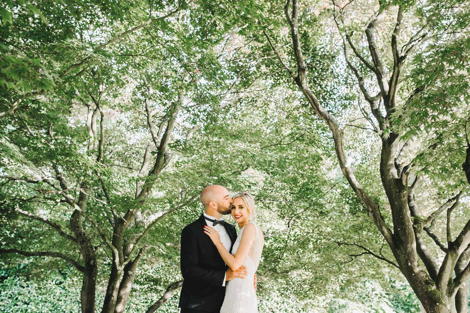 groom wearing tuxedo kisses bride surrounded by trees