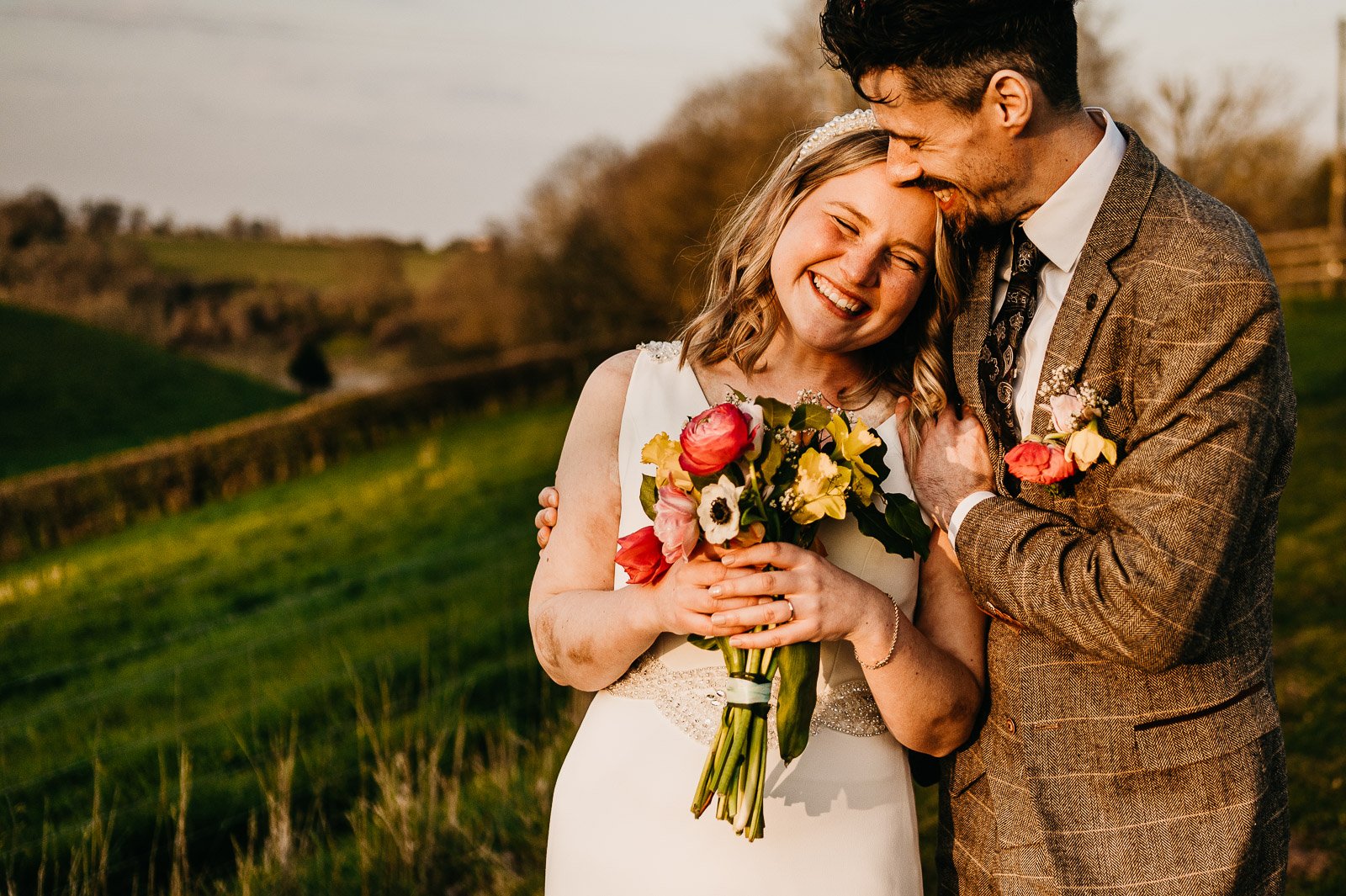 bride holds bouquet and cuddled by groom in gloucestershire countryside