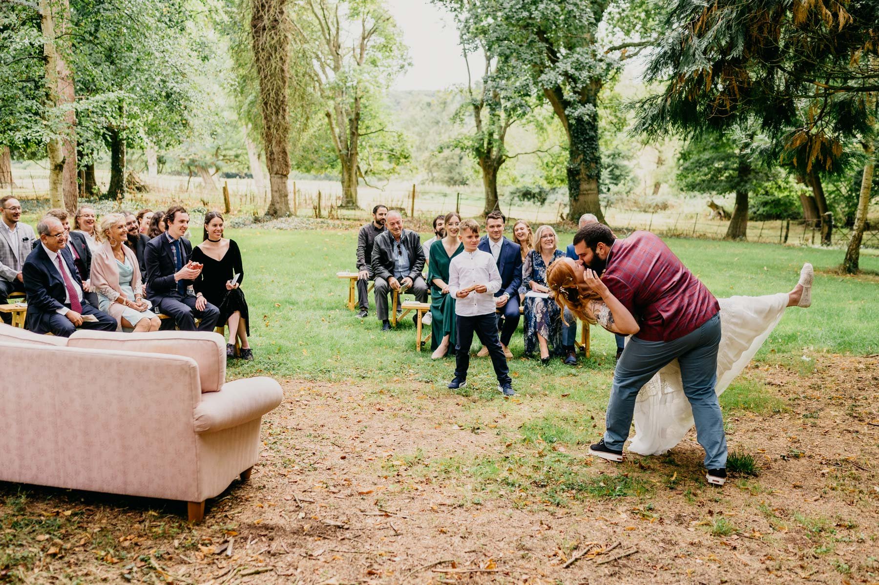 groom kisses bride during outdoor woodland wedding ceremony with sofa