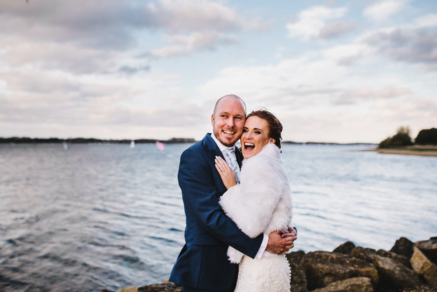bride wears white faux fur coat and is hugged by groom at rutland water