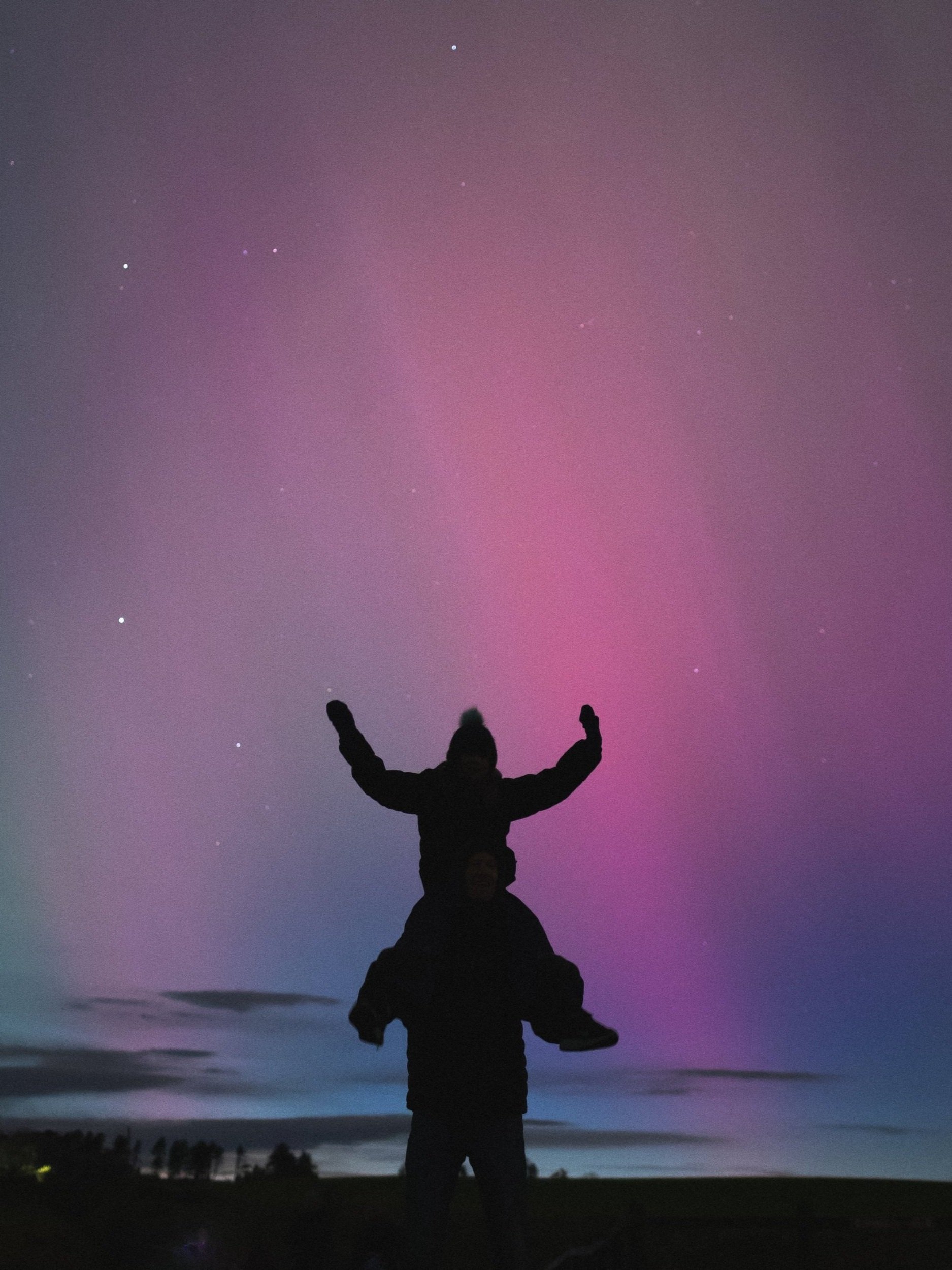 People silhouetted against a night sky with colorful aurora borealis and visible stars, with one person sitting on another's shoulders and raising their arms.