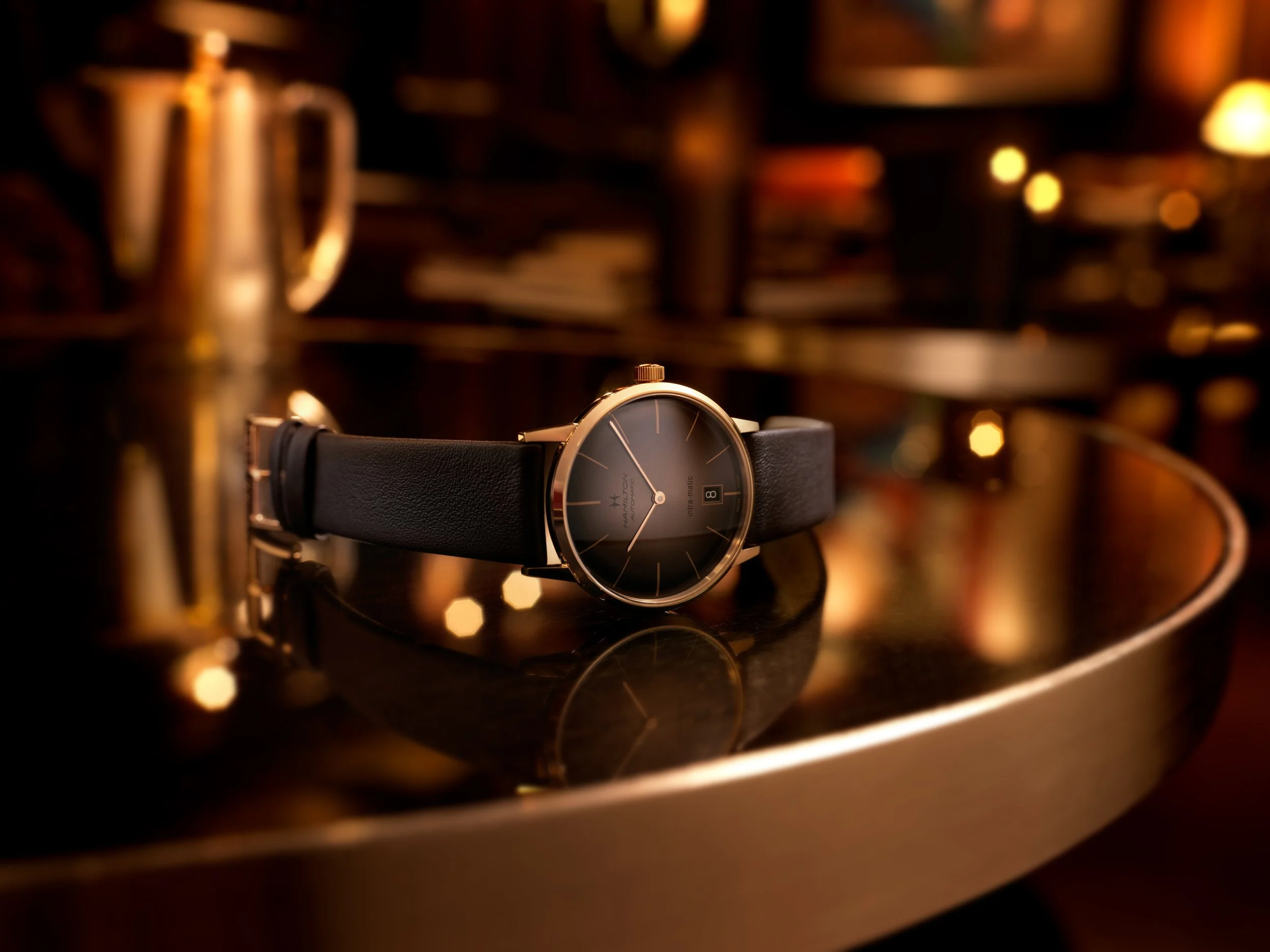 Photo publicitaire pour Hamilton: Montre noire reposant sur une surface brillante avec un fond flou, ambiance chaleureuse et raffinée.