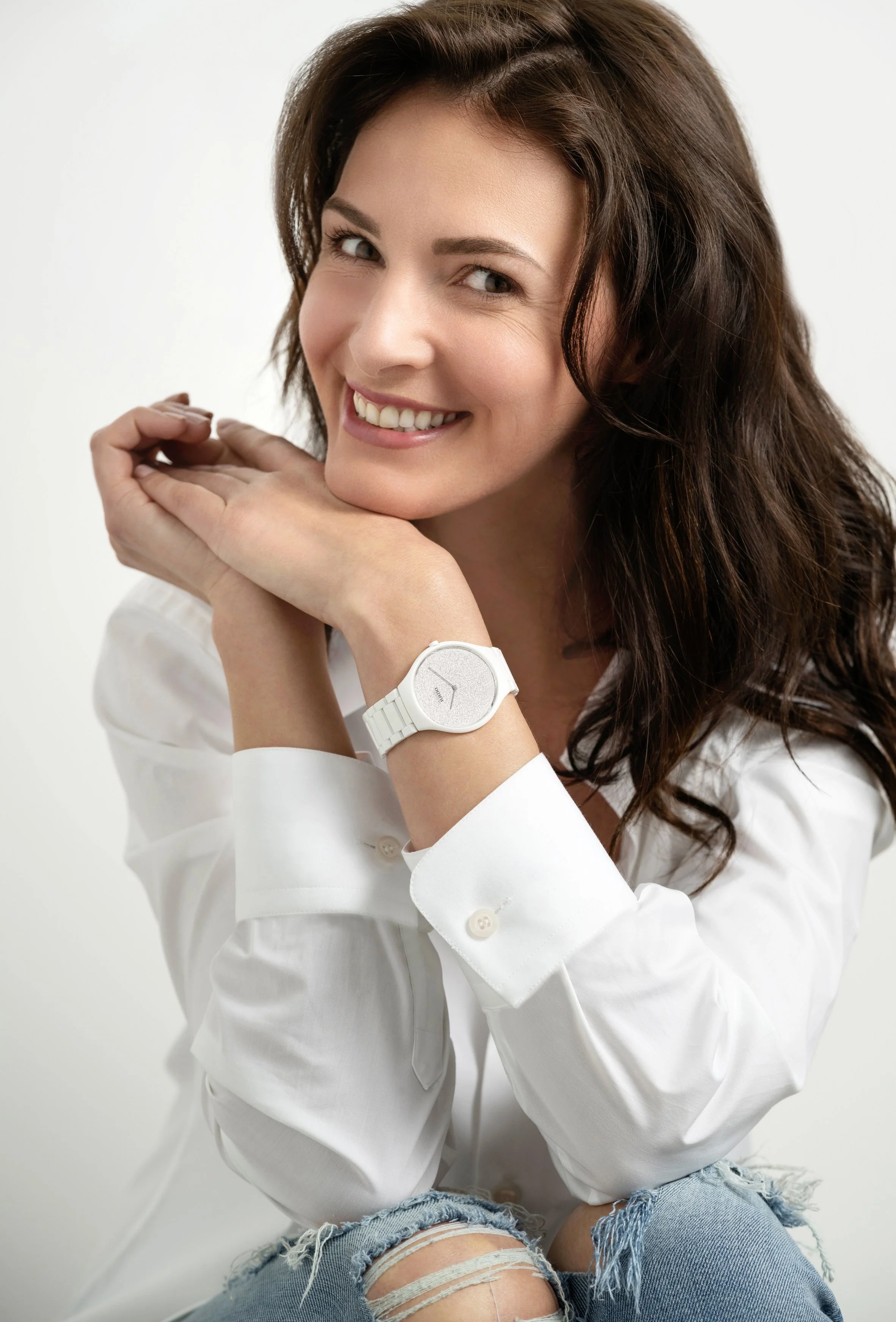 Une femme souriante avec des cheveux bruns foncés, portant une montre blanche, une chemise blanche, et un jean déchiré, posant avec les mains sous le visage.