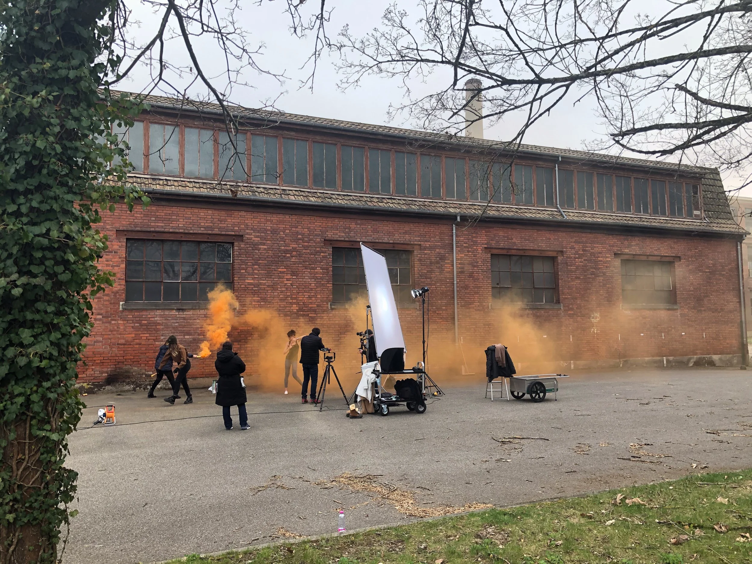 Making off shooting pro de montre: Une équipe de tournage avec des équipements professionnels, tournant une scène extérieure contre un mur en briques, avec des fumigènes orange et des personnes préparant la scène.