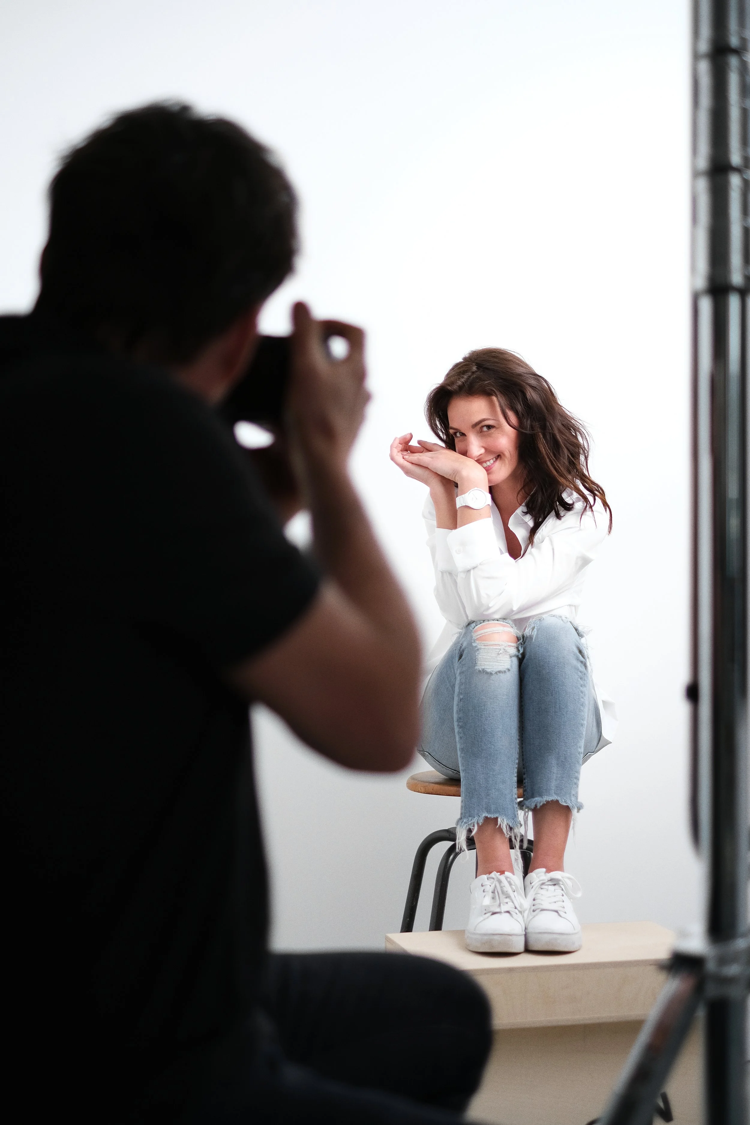 Making off shooting pro de montre: Une femme souriante en chemise blanche et jeans déchirés pose sur une caisse lors d'une séance photo, tandis qu'un photographe la photographie.