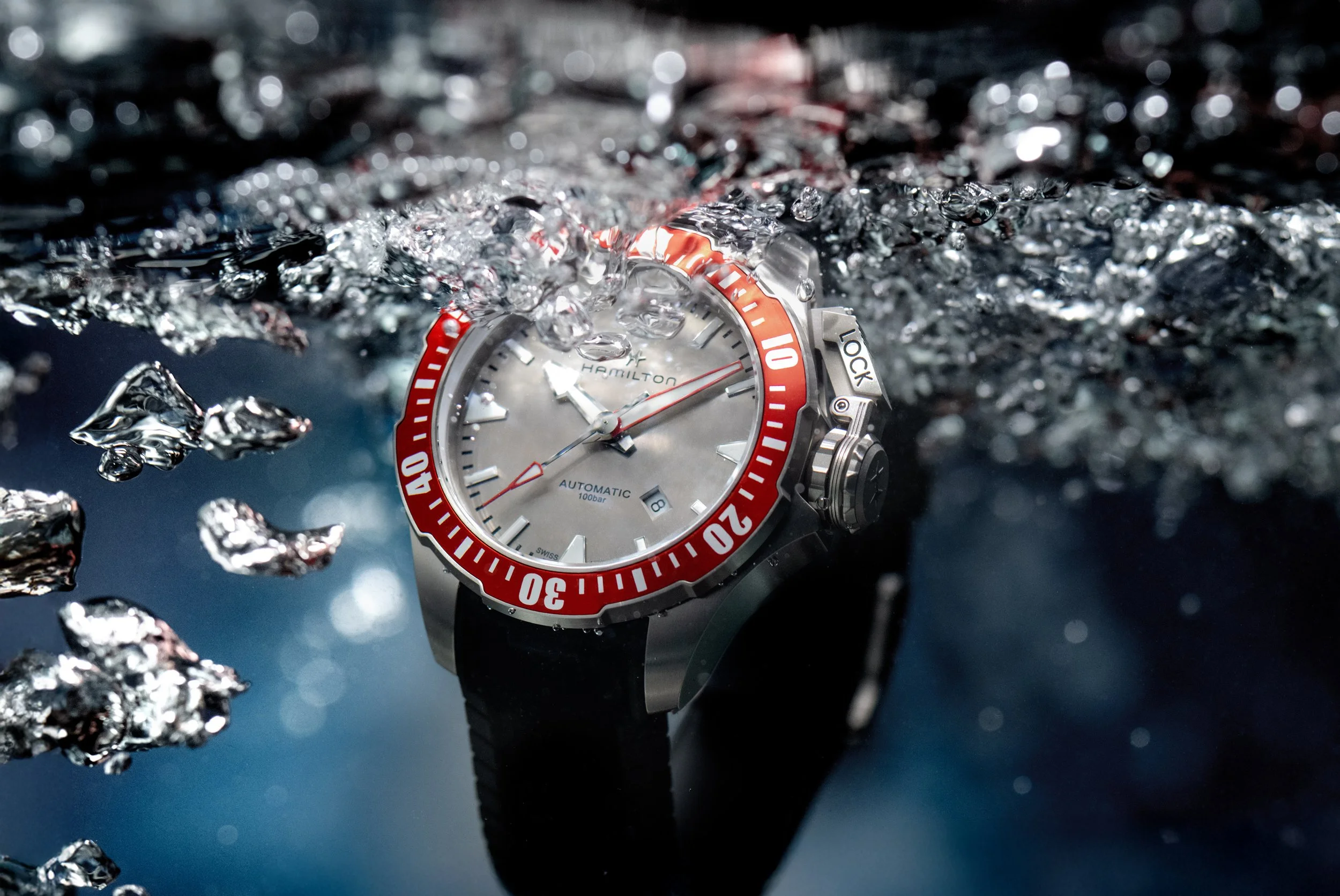 Une montre de plongée Hamilton avec un cadran blanc, un anneau rotatif rouge, immergée dans l'eau, entourée de bulles d'air.