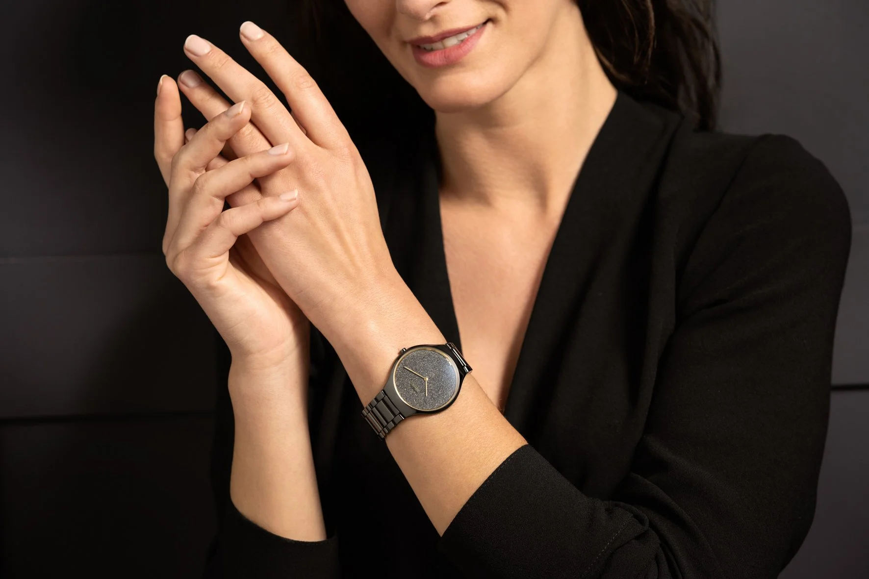 Visuel pub pour Rado : wristshot, Femme portant une montre élégante noire avec un cadran brillant et un bracelet en métal noire, tenant ses mains près de son visage, contre un fond noir.