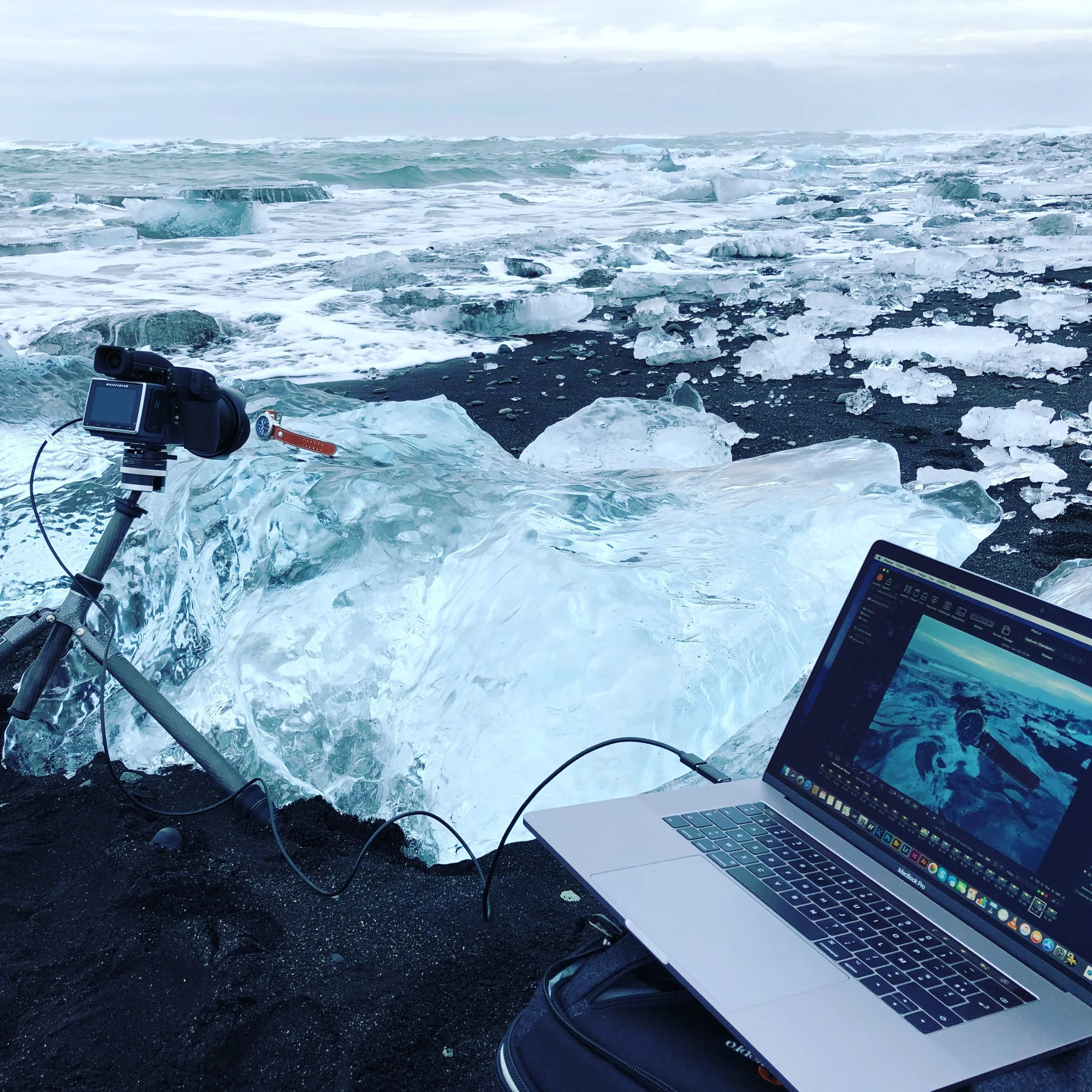Making off shooting pro de montre: Équipement de photographie, plage de sable noir couverte de morceaux de glace flottante, en arrière-plan un océan avec des icebergs et un ciel nuageux.