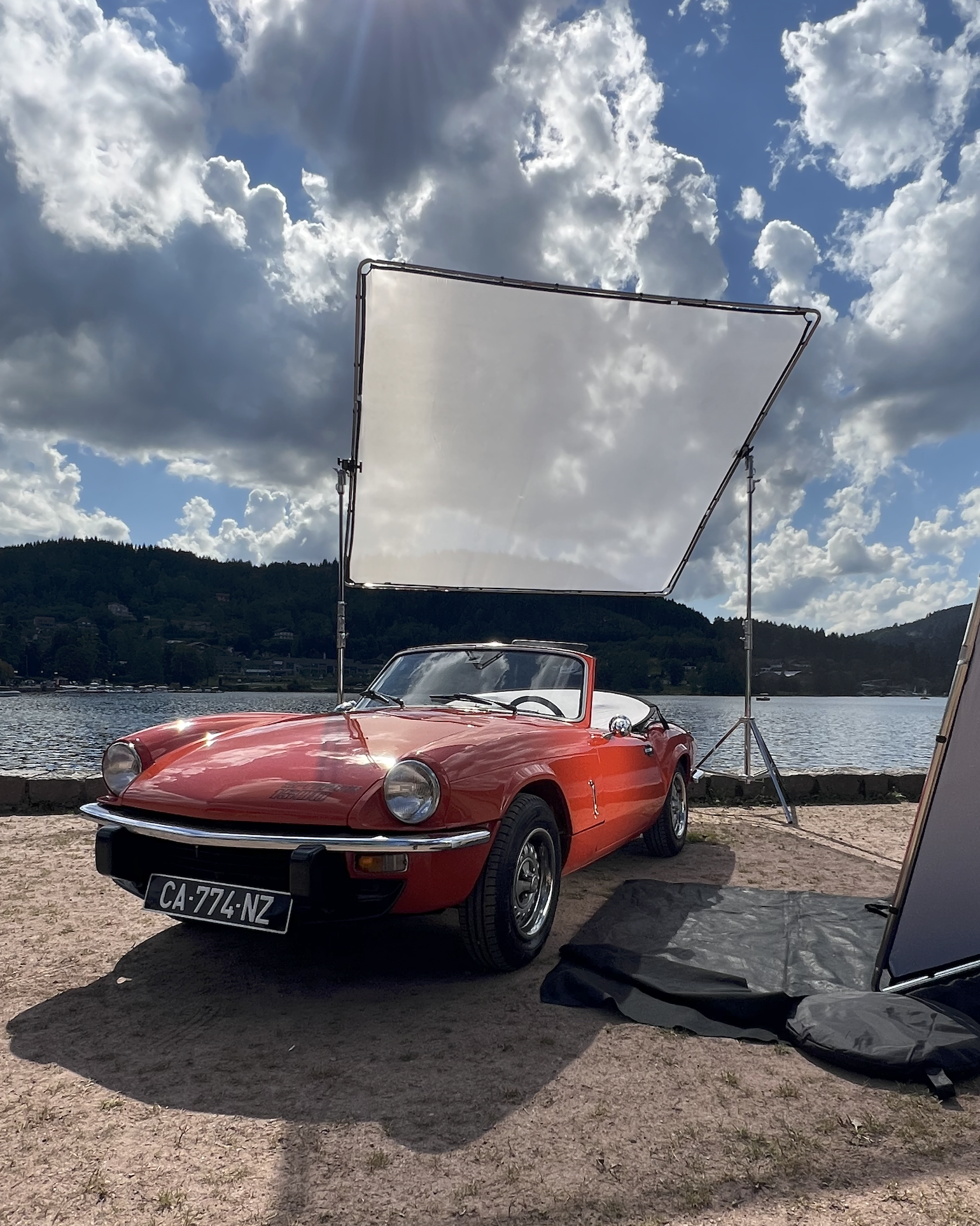 Making off shooting pro de montre: Une voiture de sport rouge près d'un lac avec un grand panneau réflecteur au-dessus, sous un ciel nuageux.